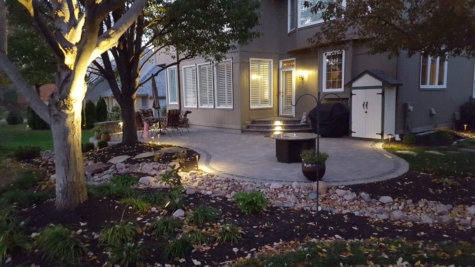 Landscaped patio and beds lit by warm landscape lighting under a large tree at dusk